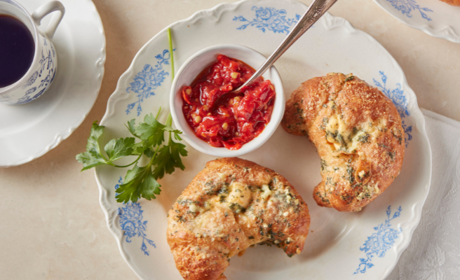Two croissants and small bowl of chili sauce on a plate with napkin and coffee cup beside.