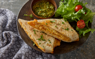 Two mac n’ cheese wraps stacked on a plate with a small side salad and small bowl of cilantro chutney sauce.