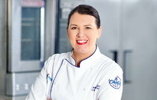 K-12 Corporate Chef, Chef Heather standing in a commercial kitchen smiling with her arms crossed and wearing a Chefs of the Mills Chef’s Coat
